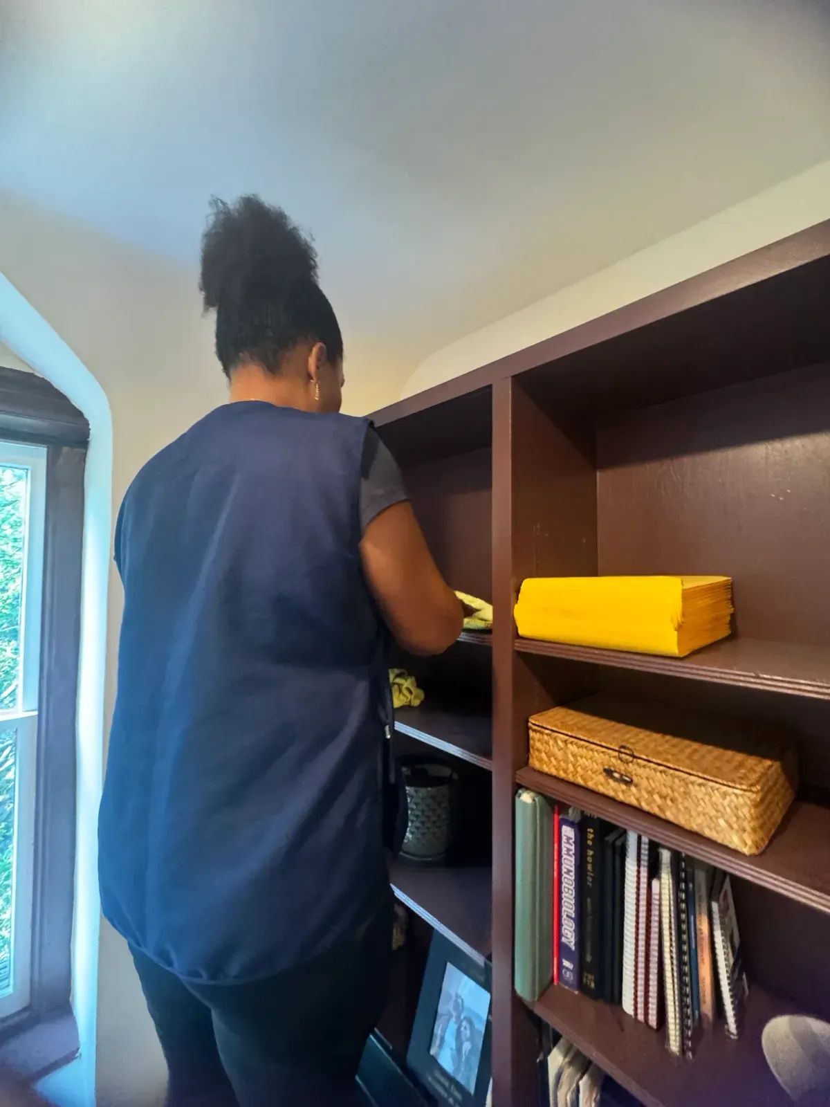 Team member wiping each shelf of the office bookcase by hand, with books and boxes set aside