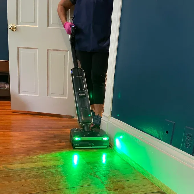A wet-dry floor washer with its green dust-detection light on, showing construction dust still on hardwood beside the baseboard