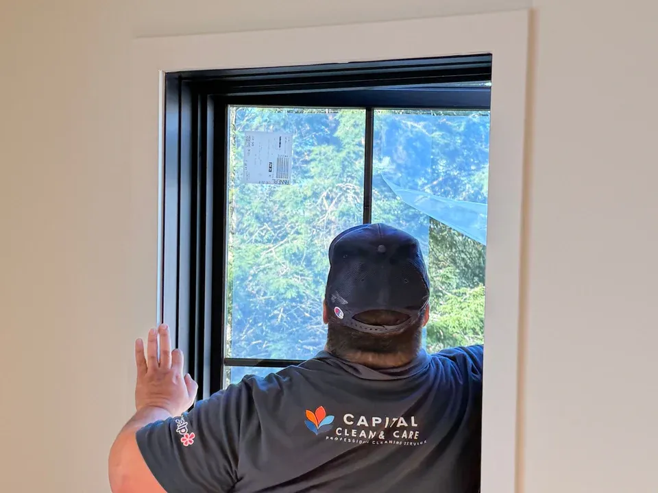 A uniformed Capital Clean Care team member cleaning a window in a newly built room, with the manufacturer's shipping label still stuck to the glass